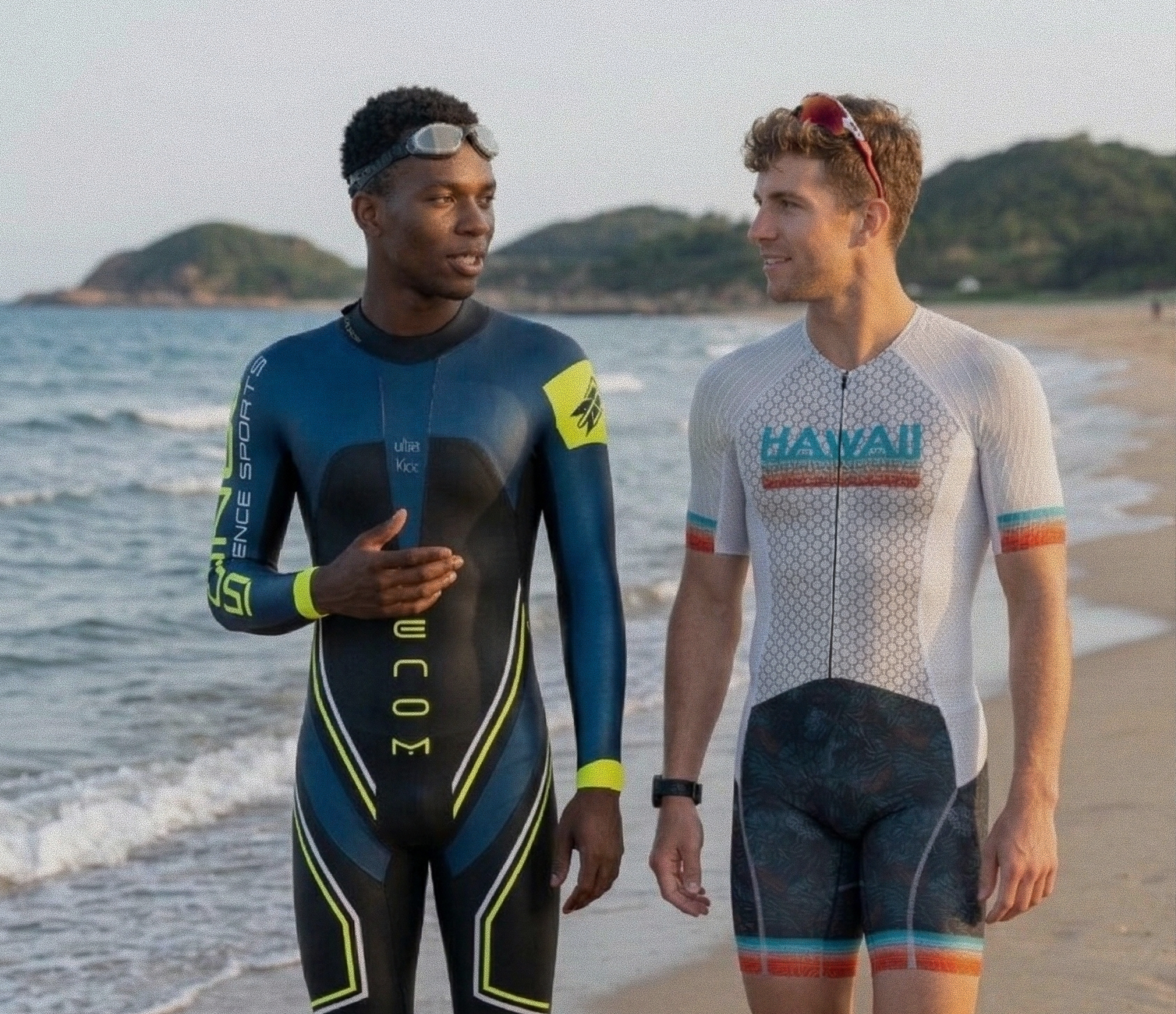 Two men on a beach: one in a full-length Venom wetsuit with blue and neon yellow details, and the other in an Elite mid-sleeve tri suit with a white top and colorful accents.